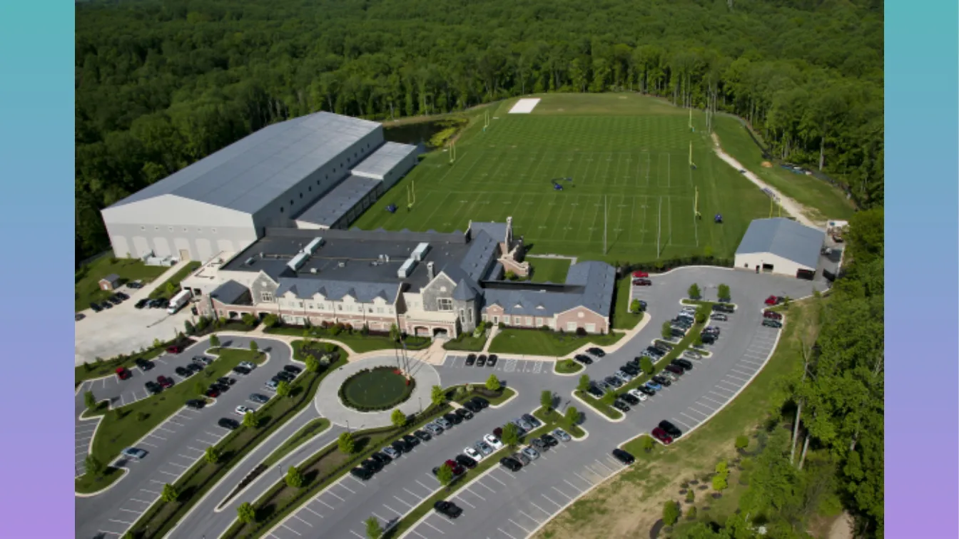 Aerial view of the Baltimore Ravens training facility in Owings Mills, Maryland, surrounded by dense forest. The image shows a large NFL complex with a curved parking lot, circular entry drive, manicured landscaping, and multiple buildings topped with a fully adhered EPDM roof system installed by Rosedale Roofing. Behind the buildings are two full-size outdoor football fields and an adjacent indoor training facility. A vertical teal-to-lavender gradient frames the left and right edges of the image.